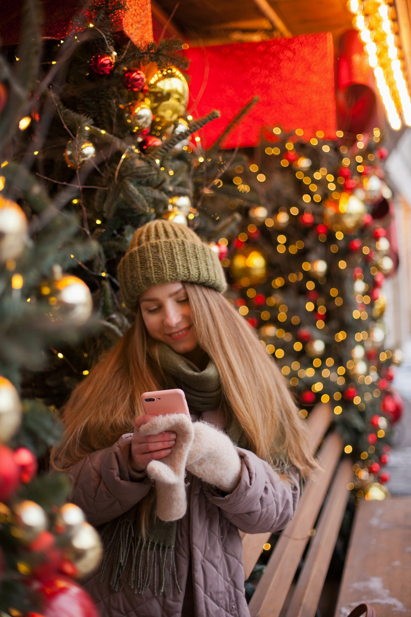 Woman using mobile phone standing near cafe or store decorated for christmas with lights and smiling